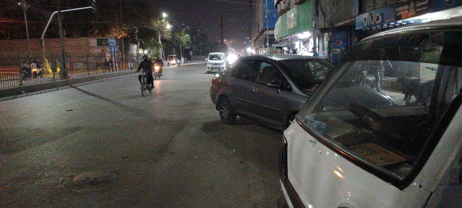“Nighttime Karachi street with glowing storefronts, vehicles, and people showing urban rhythms after dark.”