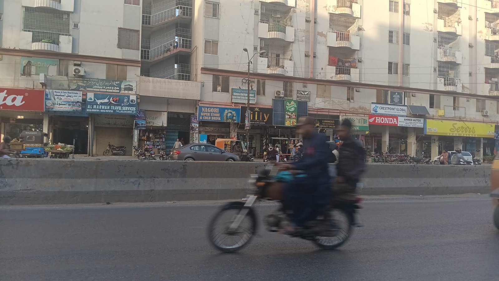 “Busy Karachi urban street with shops, motorcycles, pedestrians, and multi‑story buildings showing everyday city life.”